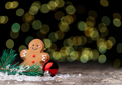 A gingerbread cookie with a pine branch and a Christmas ball on a wood table in front of a bokeh background. \nInsert your message in the background.