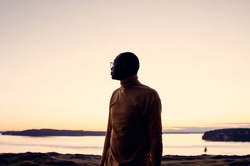A young African American looks out over a body of water at sunset, planning and dreaming of what his future holds.