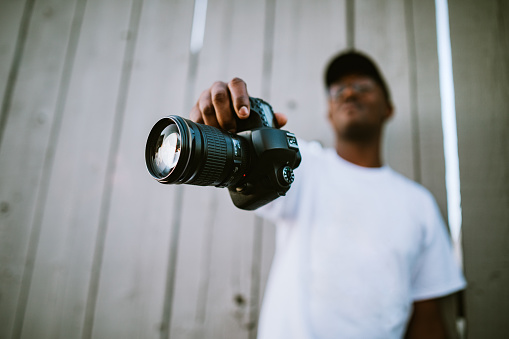 A young African American man holds out his camera and lens glass for a portrait, representing his trade of choice.