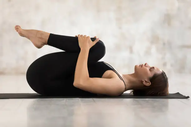 Beautiful young woman wearing black sportswear practicing yoga, relaxing in Knees to Chest pose, doing Apanasana exercise, sporty girl working out at home or in yoga studio with grey walls