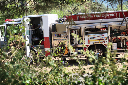 Sedona, Arizona - October 5, 2019:  A red and white fire engine from the Sedona, Arizona fire district is parked on a slight down-slope in a natural setting with doors and storage compartments open, exposing tools and other contents.  The truck is close up, filling the horizontal frame from left margin to right, with a bit of the front and rear cropped out of view.  The entire image margin is framed by foliage and pine branches such that the view of the truck is partially blocked or filtered.  No people appear in the image.