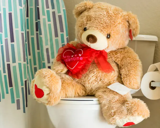 Photo of A Teddy bear in the bathroom using the toilet. Wide angle.