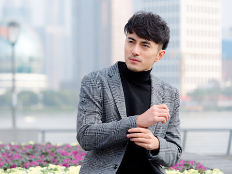 Confident and handsome Chinese young business man in casual suit sitting and looking away with hands on cuff against Shanghai bund background, Chinese businessman lifestyle concept.