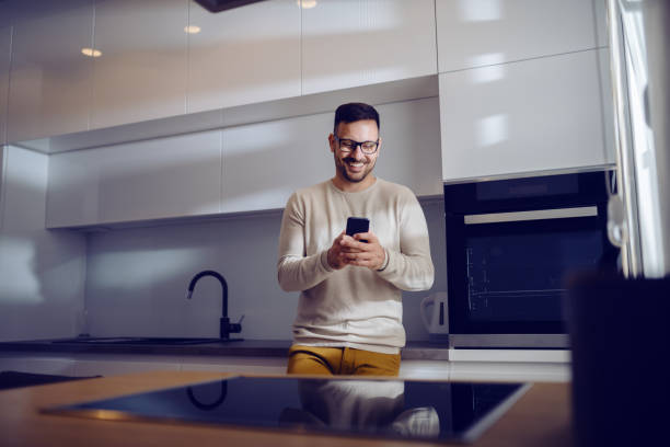 istock Attractive Caucasian smiling man dressed casual leaning on kitchen counter and using smart phone to send a message. Home interior. 1180551522