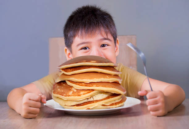 junge junge mit pfannkuchen frühstück. 7 oder 8 jahre alt glücklich und aufgeregt kind sitzt auf dem tisch essen riesigen haufen pfannkuchen lächelnd frech in zucker missbrauch und ungesunde ernährung - eierkuchen speise stock-fotos und bilder