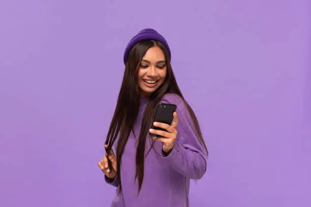 Photo of Smiling african-american girl in a hat and sweater holding smartphone isolated over purple background.