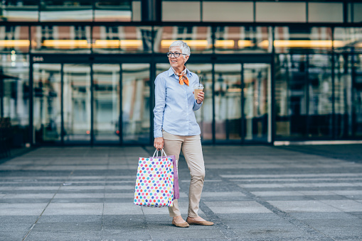 Smiling mature woman holding coffee and walking with shopping bags in the city.