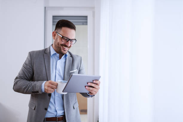 istock Smiling caucasian attractive businessman in gray suit using tablet and holding mug with coffee while standing next to window. 1180554277