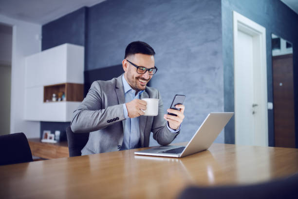 Smiling handsome Caucasian businessman in suit drinking coffee and using smart phone while sitting in dining room in morning. On desk is laptop.