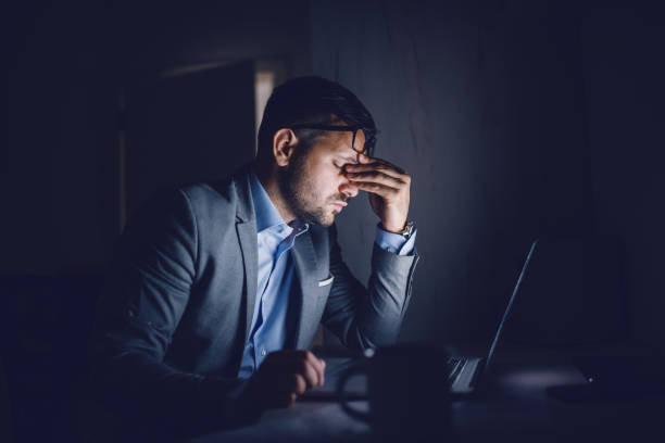 stanco duro uomo d'affari caucasico seduto in ufficio a tarda notte e strofinare gli occhi. di fronte al suo c'è il portatile. concetto di lavoro eccessivo. - lavoro straordinario foto e immagini stock