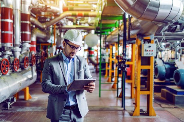 guapo gerente caucásico en traje gris y con casco en la cabeza usando tableta mientras está de pie en la planta de energía. - gasoline production fotografías e imágenes de stock