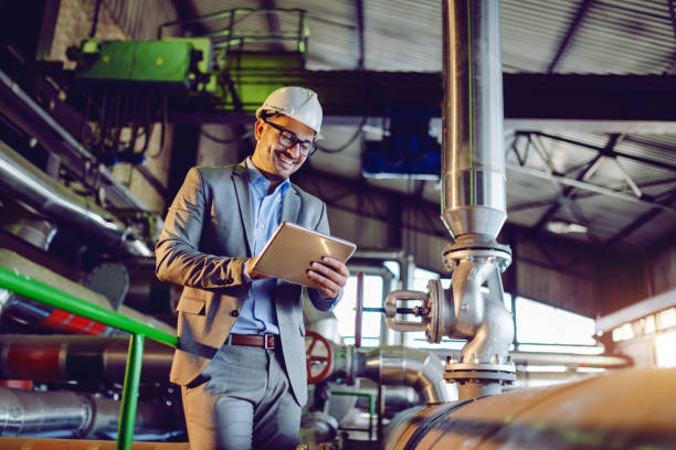 istock Handsome smiling caucasian supervisor in suit and with helmet on head using tablet while standing in power plant. 1180563305