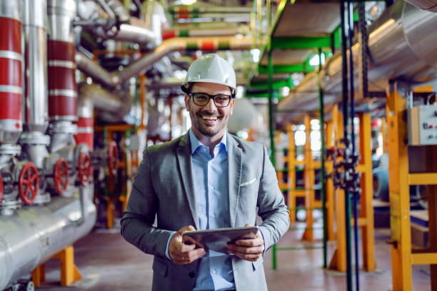istock Handsome smiling supervisor in gray suit and with white helmet on head holding tablet while looking at camera. Power plant interior. 1180563900