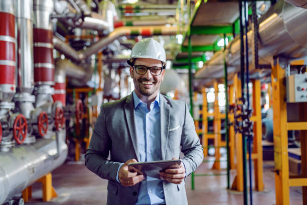 Handsome smiling supervisor in gray suit and with white helmet on head holding tablet while looking at camera. Power plant interior. Handsome smiling supervisor in gray suit and with white helmet on head holding tablet while looking at camera. Power plant interior. pipeline photos stock pictures, royalty-free photos & images