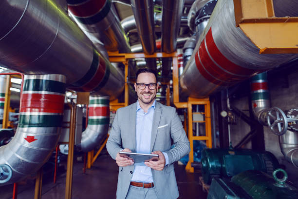 istock Handsome smiling supervisor in gray suit holding tablet while looking at camera. Energy plant interior. 1180563937