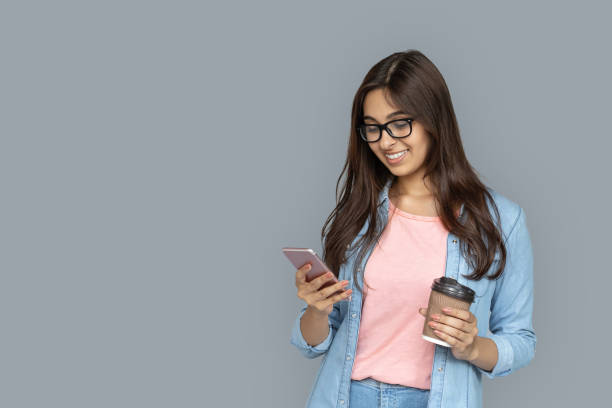 feliz sonrisa joven adulta india mujer estudiante profesional de pie aislado en el fondo gris con espacio de copia. chica mirando la pantalla del teléfono inteligente sostener taza de papel de café usando aplicaciones de medios sociales móviles - 7585 fotografías e imágenes de stock