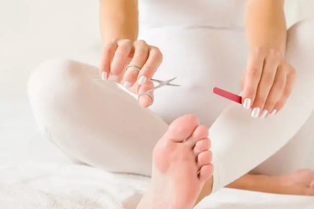 Pregnant woman in white clothes sitting on towel and trying to reach toenails, but can't to do this. Hands holding scissors and file. Uncomfortable self grooming in pregnancy time. Closeup.