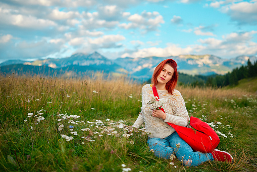 Redhead woman holding flowers sitting in the meadow in Romanian countryside, looking at camera