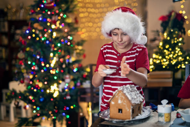 Little boy enjoying building and decorating gingerbread house for Christmas. The boy is dusting powdered sugar on the house.Nikon D850.