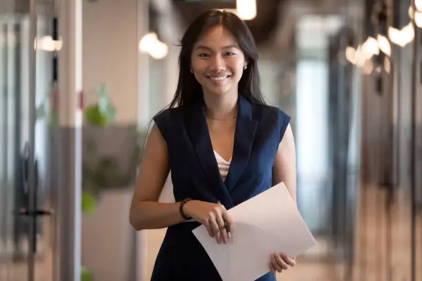 Photo of Happy asian businesswoman looking at camera stand in office hallway