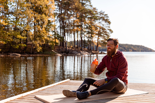 A man is sitting on an exercise mat outdoors by a lake. He is holding a protein shake.