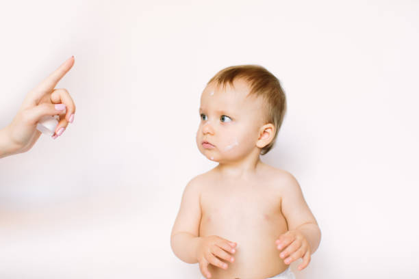 Young mother applying face kids cream on her baby girl face against light background. Hygiene, healthcare concept Young mother applying face kids cream on her baby girl face against light background. Hygiene, healthcare concept. creaming stock pictures, royalty-free photos & images