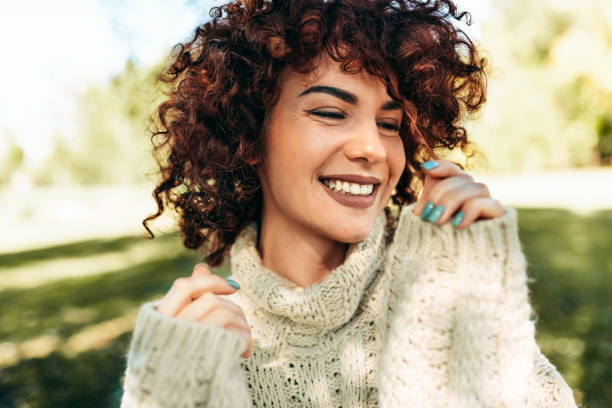 nahaufnahme porträt der schönen jungen frau lächelnd breit mit zu schüchternem lächeln, posiert vor natur hintergrund mit lockigen haaren, haben positiven ausdruck, tragen gestrickten pullover. menschen, lebensstil - women close up beautiful beauty in nature stock-fotos und bilder