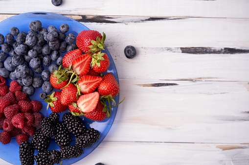 Juicy ripe organic berries of strawberries, blueberries, blackberries, raspberries on a blue plate on a light wooden background. Place for signature. Healthy eating Vegan food. Rustic background