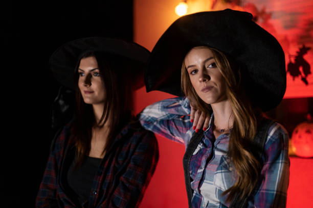 Portrait of serious beautiful young Black woman in witch hat and chiffon dress holding broom and jack-o-lantern in red room