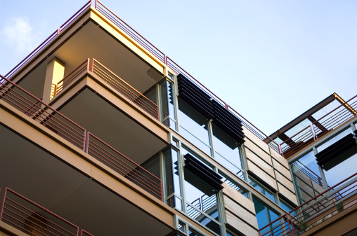 this photo is of Luxury Apartments and Condominiums in a Modern Building. the picture includes the balcony and sliding glass window at the top floor of a modern building design. the picture is an abstract background of an architectural design. the lighting is natural lighting with sunlight and blue sky is in the background. the picture was taken in a city. 