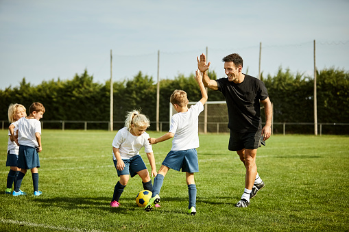 Smiling male soccer coach high-fiving with boy. Children are practicing on field. They are in sports uniforms.