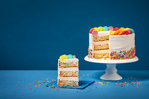 Sliced confetti Birthday cake  with rainbow colored icing and Sprinkles over a blue background.