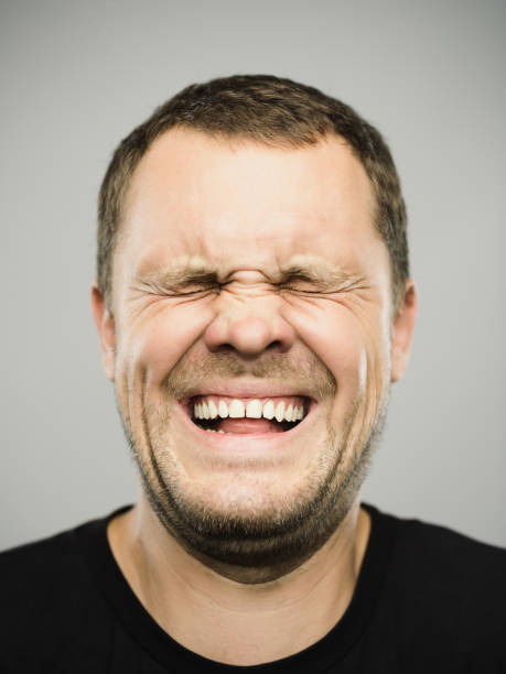 Portrait of real caucasian man with excited expression and eyes closed Close up portrait of caucasian young adult man with excited bizarre expression and eyes closed against gray white background. Vertical shot of russian real people laughing in studio with brown hair. Photography from a DSLR camera. Sharp focus on eyes. grimacing stock pictures, royalty-free photos & images