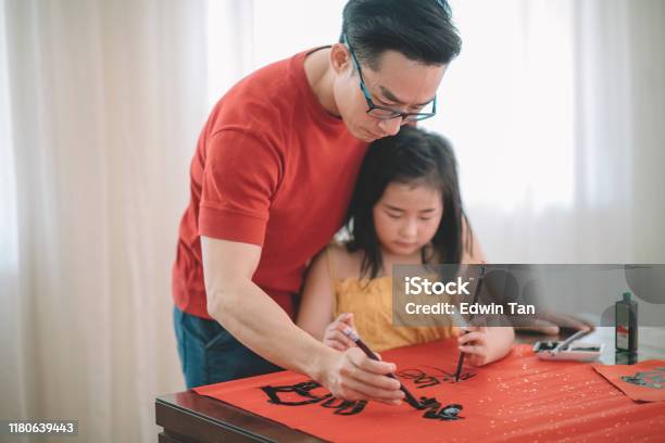 An Asian Chinese Male Practising Chinese Caligraphy For Coming Chinese New Year Celebration Home Decoration Purpose With Prosperity And Good Wording By Writing It On A Red Piece Of Paper And Teaching His Daugther Stock Photo - Download Image Now
