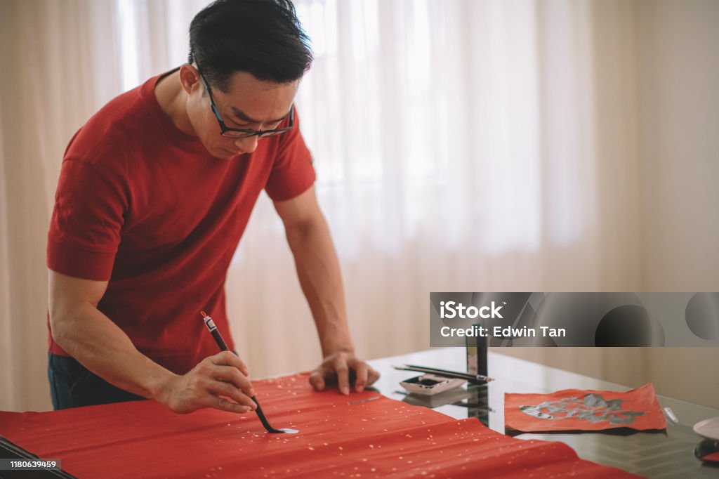an asian chinese male practising chinese caligraphy for coming chinese new year celebration home decoration purpose with prosperity and good wording by writing it on a red piece of paper Calligraphy Stock Photo