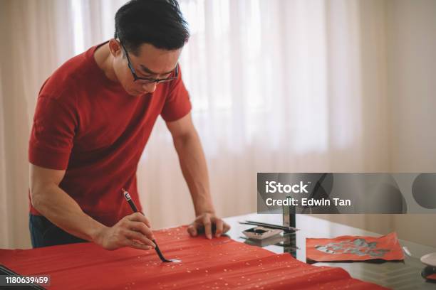 An Asian Chinese Male Practising Chinese Caligraphy For Coming Chinese New Year Celebration Home Decoration Purpose With Prosperity And Good Wording By Writing It On A Red Piece Of Paper Stock Photo - Download Image Now