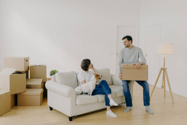 istock Positive woman and man pose in empty spacious room during relocation day, husband carries cardboard boxes with belongings, wife has telephone conversation, sits on comfortable sofa with dog. 1180651006