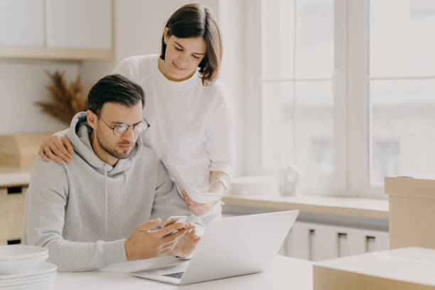 imagen de marido y mujer estudio notificación del banco, tiene teléfono móvil y papeles, trabajar en la computadora portátil, posar en la cocina durante el día de la reubicación, vestidos con ropa casual, ahorrar dinero de la familia - moving house physical activity real estate couple fotografías e imágenes de stock