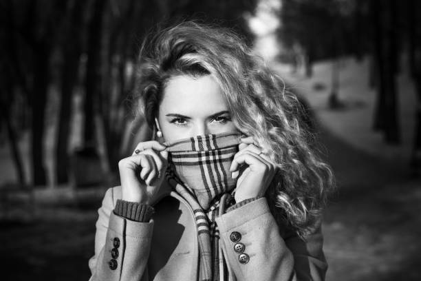 Outdoors portrait of young curly blonde woman wearing coat in winter park. Beautiful girl feeling cold and covering her face in fashionable warm scarf and looking at camera. Black and white. Caucasian female model.