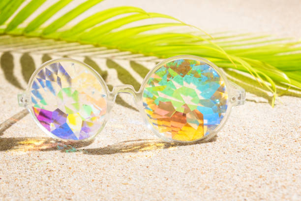 istock Funny kaleidoscope multicolour glasses on sand background with palm tree,  copy space, flat lay 1180699181
