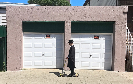New York City, USA - June 26, 2019: Hasidic Jew man riding on a kick scooter on sidewalk in Brooklyn.