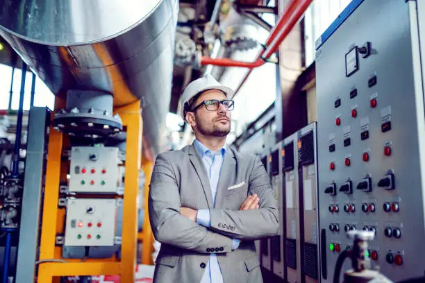 Photo of Serious handsome caucasian businessman in gray suit and with helmet on head standing with arms folded next to dashboard. Power plant interior.