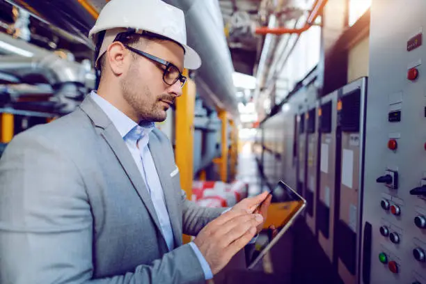 Photo of Side view of caucasian supervisor in suit and with helmet on head using tablet while standing in front of dashboard in power plant.