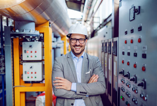 homme d'affaires caucasien de sourire dans le costume gris et avec le casque sur la tête restant avec des bras pliés à côté du tableau de bord. intérieur de centrale électrique. - energy management photos et images de collection