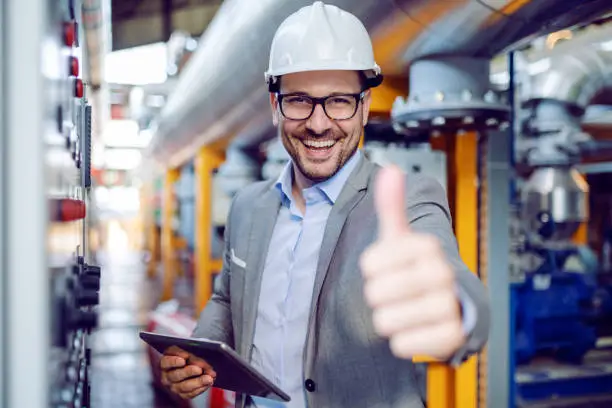 Photo of Successful caucasian supervisor in suit and with helmet holding tablet and showing thumbs up while standing next to dashboard in power plant.