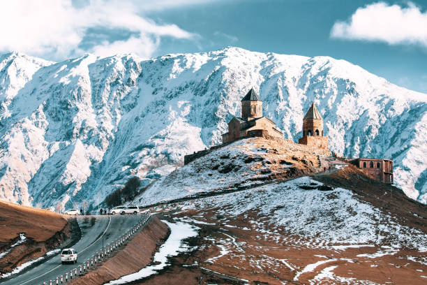 istock Stepantsminda, Georgia. Famous Gergeti Trinity Tsminda Sameba Church In Early Winter Landscape. Suv Moving Near Church In Beautiful Georgian Landscape 1180734001
