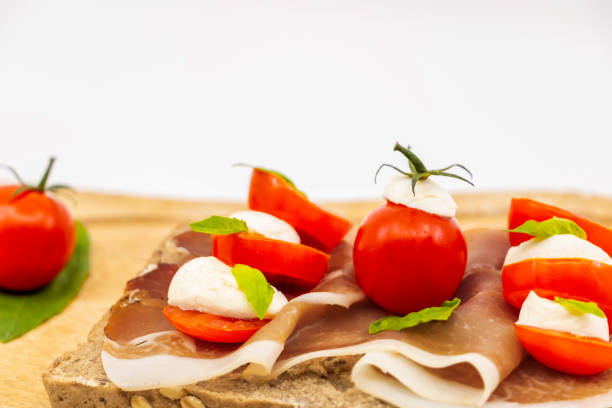 panino italiano su una fetta di pane multigrano con una fetta di prosciutto rosso da palline di pomodorini di mozzarella e basilico fresco su uno sfondo bianco isolato da un piatto di legno - mozzarella tomato sandwich picnic foto e immagini stock