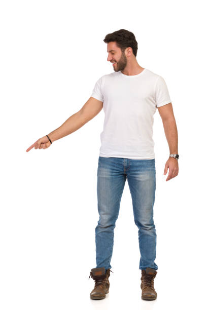 istock Smiling Young Man In Jeans And White T-shirt Is Standing And Pointing Down. Front View 1180763470