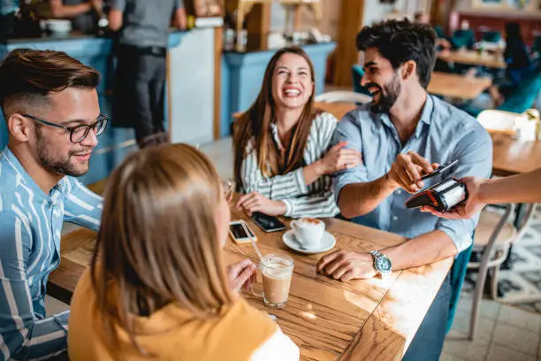 Group of friends is sitting in a coffee shop and a guy is paying bill with a smart phone