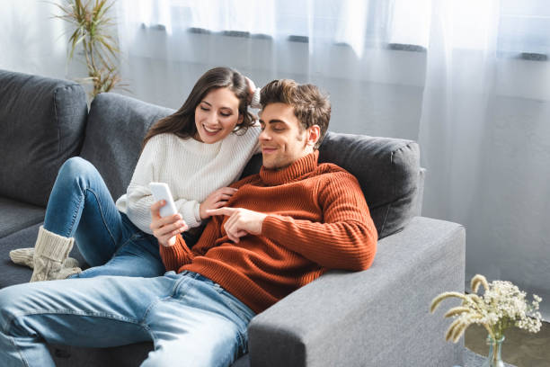 smiling girlfriend and boyfriend in sweaters looking at smartphone smiling girlfriend and boyfriend in sweaters looking at smartphone house phone stock pictures, royalty-free photos & images